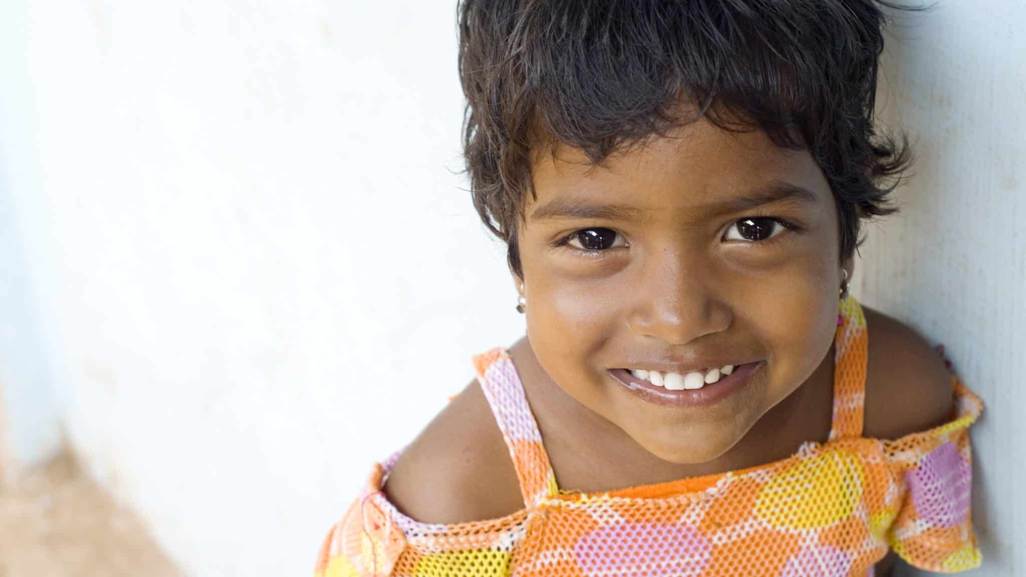 Smiling young girl in India in a bright orange and pink dress, looking up at the camera.